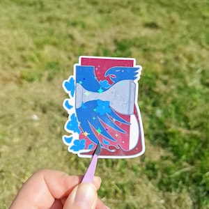 May include: A sticker featuring a blue and red design of a bird inside a bottle. The bird has a stylized design with blue and white accents. The sticker is held by a person's hand with a pair of pink tweezers.