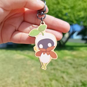 May include: A wooden keychain featuring a cartoon-style character with a white body, black face, and brown accents. The keychain has a small green leaf and a metal clasp for attachment. The background is a blurred outdoor scene.