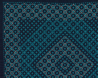 May include: A dark blue fabric square with a geometric pattern embroidered in light blue and white thread.