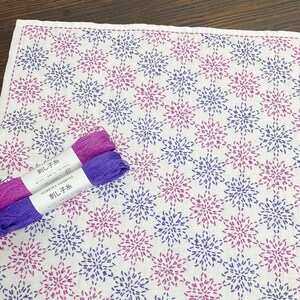 May include: White fabric with a repeating pattern of pink and purple floral designs. Two spools of thread, one pink and one purple, are placed on the fabric. The fabric has a stitched border and the thread spools have Japanese text.