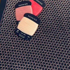 May include: A navy blue fabric square with Sashiko stitching in a geometric pattern using pink thread. Three spools of Sashiko thread in pink and peach tones are placed on the fabric. The spools have black labels with white text.