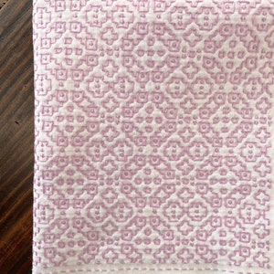 May include: A white linen tea towel with a delicate purple geometric pattern embroidered on it. The towel has a subtle texture and a simple, elegant design.