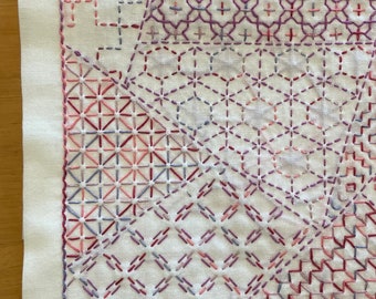 May include: A white fabric square with intricate embroidery designs in shades of pink, red, and purple. The embroidery patterns are geometric and create a visually appealing patchwork effect.