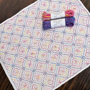 以下が含まれることがあります： カラフルな刺繍の幾何学模様が施された白い生地。ピンク、黄色、青色のキラキラした刺し子糸が2つ上にあります。生地には白い縁取りがあり、濃い木材の表面に置かれています。