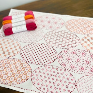 以下が含まれることがあります： ピンクとオレンジの色合いで刺繍された楕円形の模様が施された白い生地。色を合わせた色の糸がいくつか生地の上に置かれており、手芸や裁縫のプロジェクトを示唆しています。デザインは、さまざまな幾何学模様と花柄を特徴としています。