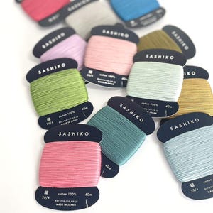 May include: A selection of Sashiko thread spools in a range of colours, such as pink, green, blue, and grey. Each spool is labelled with "Sashiko" and thread details, with a length of 40m. The spools are displayed on a white background.