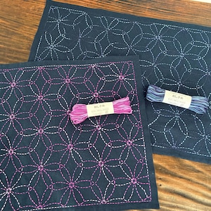 Op de afbeelding: Marineblau geborduurde placemats met een bloemmotief. De placemats hebben witte en roze stiksels. Twee spoelen garen, één roze en één blauw, zijn inbegrepen. De placemats liggen op een houten oppervlak.