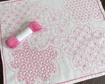 Può includere: Tessuto bianco con ricamo floreale rosa e una bobina di filo rosa abbinata. Il ricamo presenta motivi intricati e un bordo cucito. La bobina di filo ha testo giapponese.