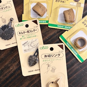 以下が含まれることがあります： Cloverブランドの裁縫道具セット。パッケージ入りのベージュのレザー指ぬき、ダークメタルの円形糸切りペンダント、ハート型の針通し、日本語のテキストが記載されたゴールドカラーの糸切りリングが含まれています。