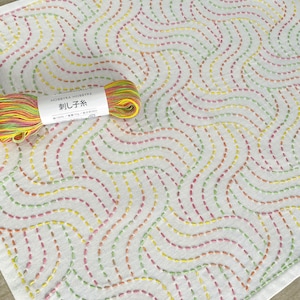 May include: A white fabric square with a repeating pattern of colorful stitched lines. The fabric is decorated with a wave-like design. A skein of multi-colored embroidery floss is included. The floss is labeled "Sashiko" in Japanese characters.