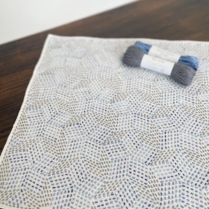 May include: A white fabric square with a geometric pattern of blue and gray embroidery stitches. Two skeins of blue embroidery floss are on top of the fabric.