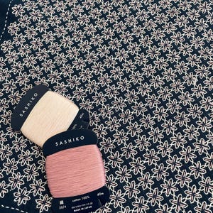 May include: Two spools of pink and white Sashiko thread, labelled "Sashiko" and "cotton 100%", on a dark blue fabric with a white and pink embroidered pattern. The fabric is 40 cm wide.