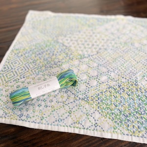 Può includere: Un quadrato di tessuto bianco con un ricamo verde, blu e giallo. Un gomitolo di filo da ricamo verde, blu e giallo è sopra il tessuto. L'etichetta del gomitolo di filo dice "HOWRTRA HOUSE 刺し子糸".