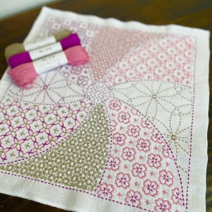 以下が含まれることがあります： ピンクとベージュの色合いで幾何学模様と花柄が刺繍された白い正方形の織物。ピンクとベージュの糸が2つ上に置かれています。この織物は、手芸や装飾用に使用される可能性があります。