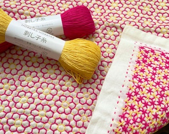May include: A white cotton dishcloth with a pink and yellow geometric pattern. Two spools of embroidery floss, one pink and one yellow, are included. The text on the spools reads "刺し子糸" which translates to "Sashiko thread".