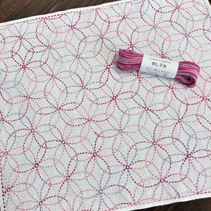May include: A white fabric cloth featuring a repeating geometric circle pattern stitched in variegated pink, purple, and blue thread. A skein of matching variegated embroidery thread with a label reading HOBBYRA HOBBYRE is placed on top.