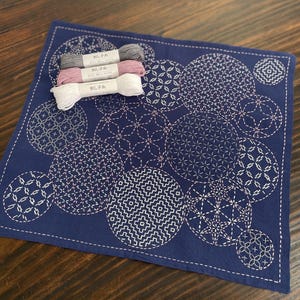 Può includere: Un tessuto quadrato blu navy con motivi circolari ricamati in bianco. Tre rocchetti di filo grigio, rosa e bianco sono sopra. Il testo giapponese sui rocchetti di filo è parzialmente visibile.