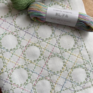 Könnte beinhalten: Zwei Spulen Stickgarn, eine grün und eine mit einem Regenbogen an Farben, mit dem Text "刺し子糸" (Sashiko-Garn) und "綿100%/重量10g/長さ約85m" (100% Baumwolle/Gewicht 10g/Länge ca. 85m) auf den Etiketten. Die Spulen liegen auf einem weißen Stoff mit einem Rautenmuster, das mit grünem, rosa, blauem und gelbem Garn gestickt ist.