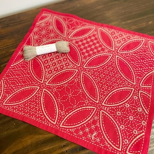 May include: A red square textile with white embroidered floral patterns. A spool of beige thread rests on the textile. The textile features a repeating design of large, stylized flower shapes and geometric patterns.