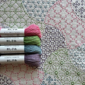 Könnte beinhalten: Vier Stickgarnstränge in Pink, Grün, Blau und Lila, jeweils mit einem weißen Papieretikett mit der Aufschrift HOBBYRA HOBBYRE und japanischem Text, auf einem weißen Stoffhintergrund mit einem geometrischen Muster in passenden Farben.