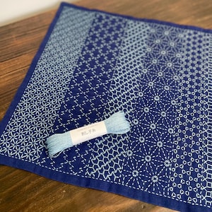 Könnte beinhalten: Ein marineblaues und weißes Textil mit geometrischem Muster. Eine hellblaue Garnrolle mit japanischem Text liegt auf dem Textil. Das Textil weist ein Farbverlaufsmuster auf, das von hellblau zu dunkelblau übergeht.