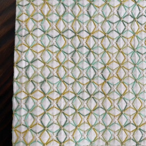 May include: Close-up of a white fabric with a geometric pattern of embroidered diamonds. The embroidery uses light green, yellow-green, and cream-colored thread. The fabric has a textured appearance and a decorative border.