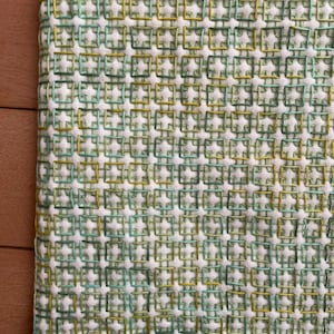 以下が含まれることがあります： 緑と黄色の色合いで刺繍された、繰り返しの正方形のパターンが特徴の白い生地のクローズアップ。 刺繍はテクスチャード加工された表面を作り出し、下端に沿って緑色のステッチの境界線があります。 この生地はおそらく装飾的なテキスタイルです。