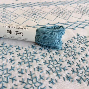 May include: A skein of blue embroidery thread labeled HOBBYRA HOBBYRE with 100% cotton, 10g, and 85m text, resting on white fabric featuring blue geometric and snowflake-like embroidery patterns stitched in a repeating, symmetrical design.
