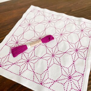 May include: A white cotton tea towel with a geometric pattern of pink stars stitched on it. The towel is accompanied by a spool of pink embroidery floss.