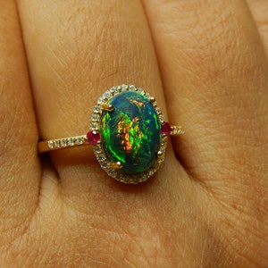 May include: A gold ring featuring an oval-shaped black opal with vibrant green and orange flashes. The center stone is surrounded by a halo of small white diamonds, with a single small red gemstone on each side of the band, which is set with diamonds.