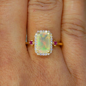 Pode incluir: Um anel de ouro com uma pedra central de opala retangular rodeada por um halo de pequenos diamantes brancos. O anel inclui dois pequenos rubis vermelhos redondos cravados em cada lado da opala central, numa banda de ouro polido.