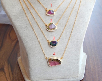 May include: Four gold necklaces with gemstone pendants. Each necklace features a different colored gemstone, including pink, purple, and green, set in a gold frame with a halo of small, clear stones. The necklaces are displayed on a white mannequin.