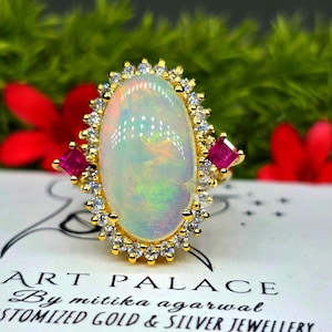Puede incluir: Un anillo de oro con una gran piedra de ópalo ovalada en el centro rodeada por un halo de pequeñas piedras claras y dos piedras rosas cuadradas a los lados. El anillo se muestra sobre una tarjeta blanca con el texto ART PALACE By mitika agarwal.