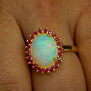 May include: A gold ring featuring an oval opal gemstone with a halo of small, round, red gemstones. The opal has a milky white base with hints of green and blue. The ring is set on a gold band.