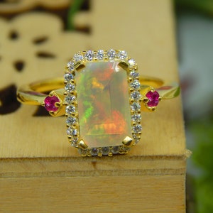 Könnte beinhalten: Ein goldener Ring mit einem großen, rechteckigen Opal mit einem Kranz aus kleinen, klaren Edelsteinen. Zwei kleine, runde, rote Edelsteine flankieren den Opal. Der Ring liegt auf einer Holzoberfläche.