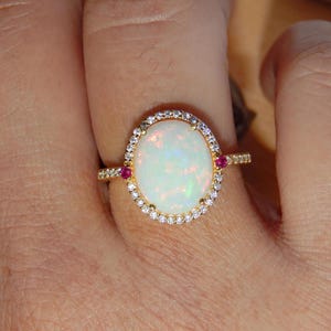 Puede incluir: Un anillo de oro con una gran piedra central de ópalo blanco en forma ovalada rodeada por un halo de pequeños diamantes transparentes. Dos pequeños rubíes rojos redondos están engastados en la banda a los lados del ópalo central, y la banda está decorada con diminutos diamantes.
