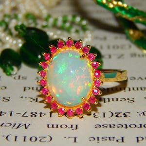 May include: A gold ring with an oval opal, encircled by small pink gemstones. The opal displays iridescent colours. The ring is set on a gold band. The ring is displayed on a printed surface.