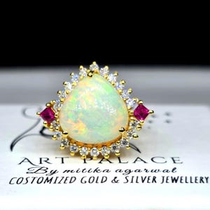 May include: A gold ring featuring a large pear-shaped iridescent opal center stone, surrounded by a halo of small clear white stones and two square-cut pink stones on the sides. The ring rests on a white card labeled ART PALACE By mitika agarwal.