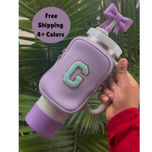 May include: A purple insulated water bottle with a handle and a zippered pouch. The bottle has a light blue letter "G" with gold glitter on the front. The bottle has a purple bow on top.