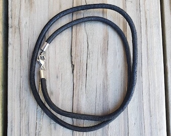 Pode incluir: Um cordão de colar preto com fecho prateado, sobre uma superfície de madeira envelhecida. O cordão é feito de um material liso e escuro e está em forma de laço. O fecho é de design metálico simples.