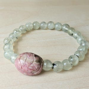 Op de afbeelding: Een armband met lichtgroene en heldere kralen met zwarte insluitsels. De armband heeft een centrale, ovaalvormige roze steen met gesneden geometrische patronen. De armband wordt getoond op een licht houten oppervlak.