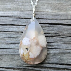 May include: A teardrop-shaped pendant necklace featuring a polished, translucent stone with white and peach-colored inclusions. The stone is suspended from a delicate silver chain. The pendant has a unique, natural pattern and a small cavity.