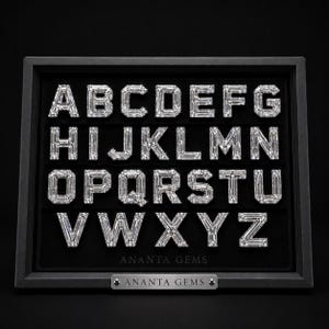 Może przedstawiać: Czarne pudełko ekspozycyjne zawierające pełny alfabet od A do Z, gdzie każda litera uformowana jest z przezroczystych kamieni szlachetnych o prostokątnym szlifie. Napis ANANTA GEMS znajduje się pod literami oraz na srebrnej metalowej tabliczce.
