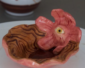Puede incluir: Un plato de cerámica con un diseño en forma de flor. El plato tiene un borde rosa y un interior marrón, similar a la veta de la madera. Una flor rosa con un ojo en el centro está unida al plato. Un jarrón blanco está en el fondo.