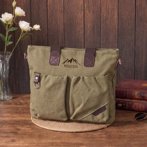 May include: An olive green canvas tote bag featuring two front pockets, dark brown fabric handles, and a mountain graphic with the text WESTEN printed above the pockets. The bag includes a silver metal ring attachment on the left front pocket.