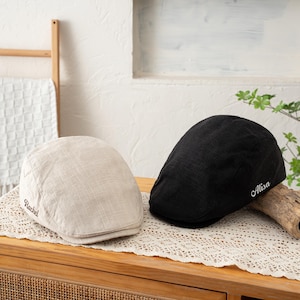 May include: A beige flat cap with the name Rachel embroidered in brown and a black flat cap with the name Alisa embroidered in white. Both caps feature a textured fabric construction with a rounded crown and a short, stiff front brim.