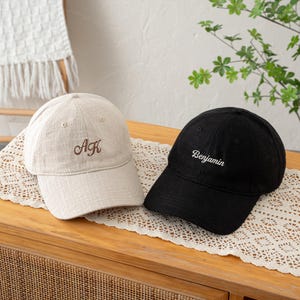 May include: A beige baseball cap with the embroidered brown letters AK and a black baseball cap with the embroidered white name Benjamin. Both caps feature a textured fabric, a curved brim, and a button on the top center of the crown.