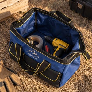 May include: A blue and black tool bag with yellow stitching, featuring the word Austin and a mountain graphic on the front. The open bag contains a yellow cordless drill, a roll of silver tape, and a screwdriver, with black handles attached.