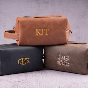 May include: Three leather toiletry bags in brown, black, and dark brown. The brown bag has the embroidered letters "KIT", the black bag has "GEK", and the dark brown bag has "MATTHEW GROOMSMEN" with a deer antler design. Each bag has a zipper closure and a handle.