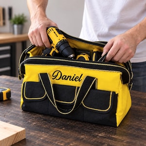 May include: A yellow and black tool bag featuring the name Daniel embroidered in black script on the front. The bag has multiple exterior pockets, black fabric handles, and a wide zippered opening containing a yellow and black power drill.